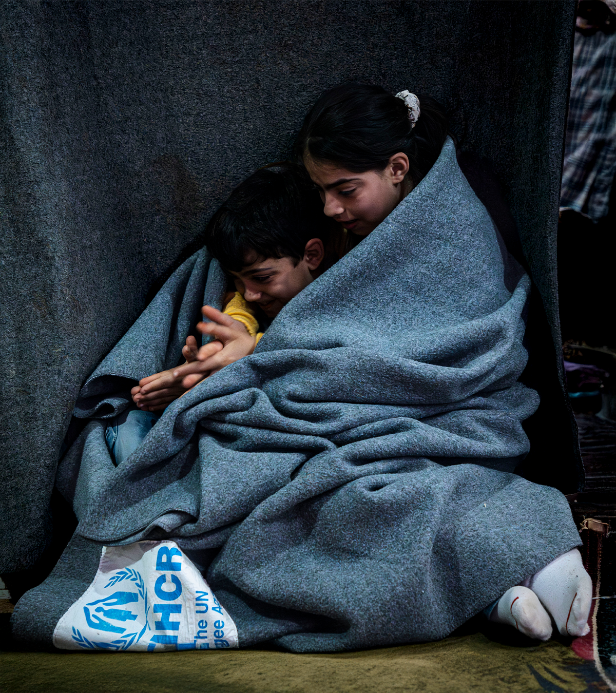 winterLP-image Duas crianças abraçadas envolvidas em um cobertor do ACNUR. Elas estão sorrindo e brincando com as mãos.